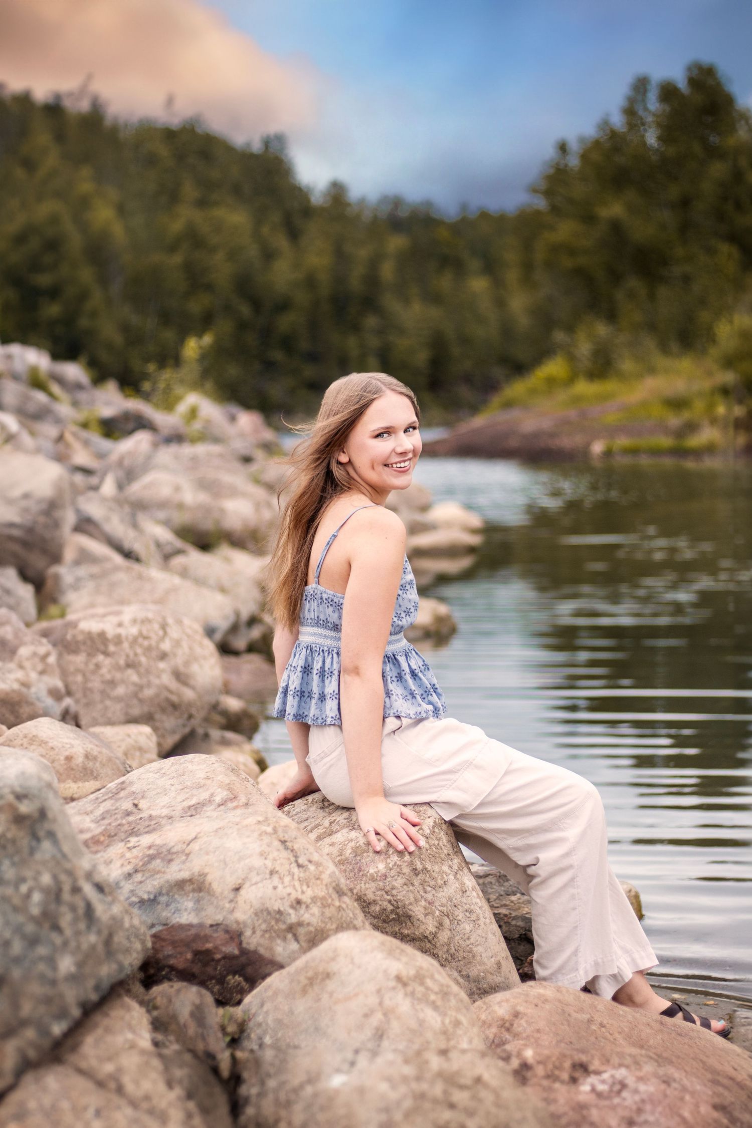 Someone in a blue and white sleeveless top and white pants sits on rocky shoreline by tranquil lake surrounded by forest.