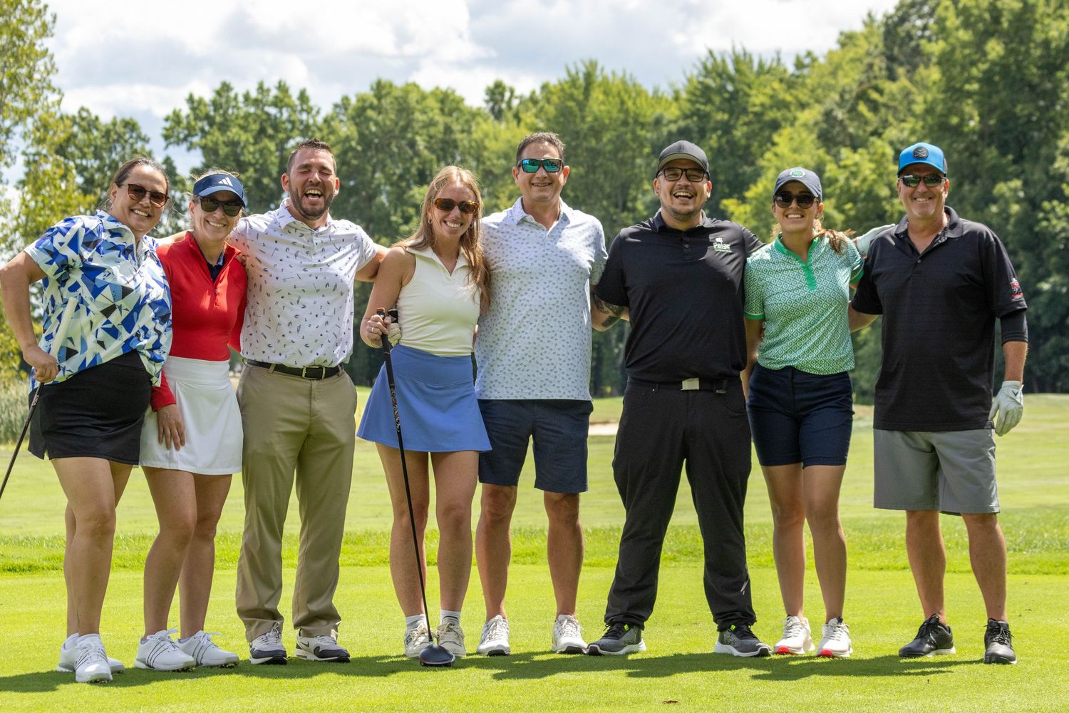 A group of eight smiling golfers posing together on a sunny golf course fairway.