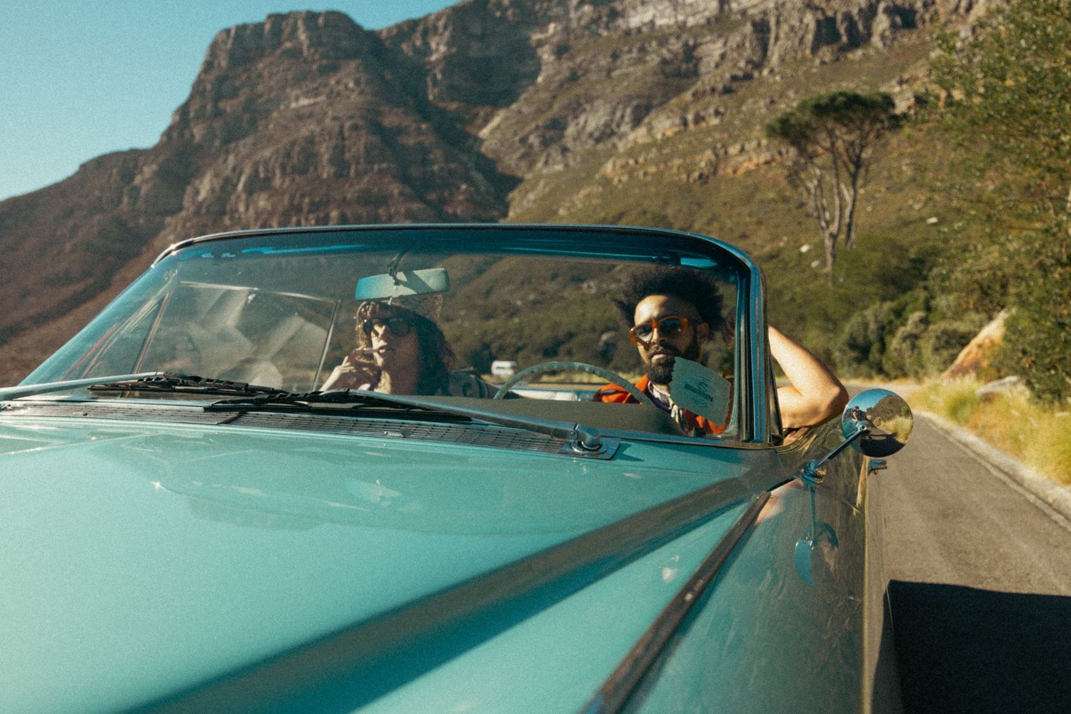 A turquoise classic car drives along a scenic mountain road on a Cape Town photo production with rugged cliffs in the background.