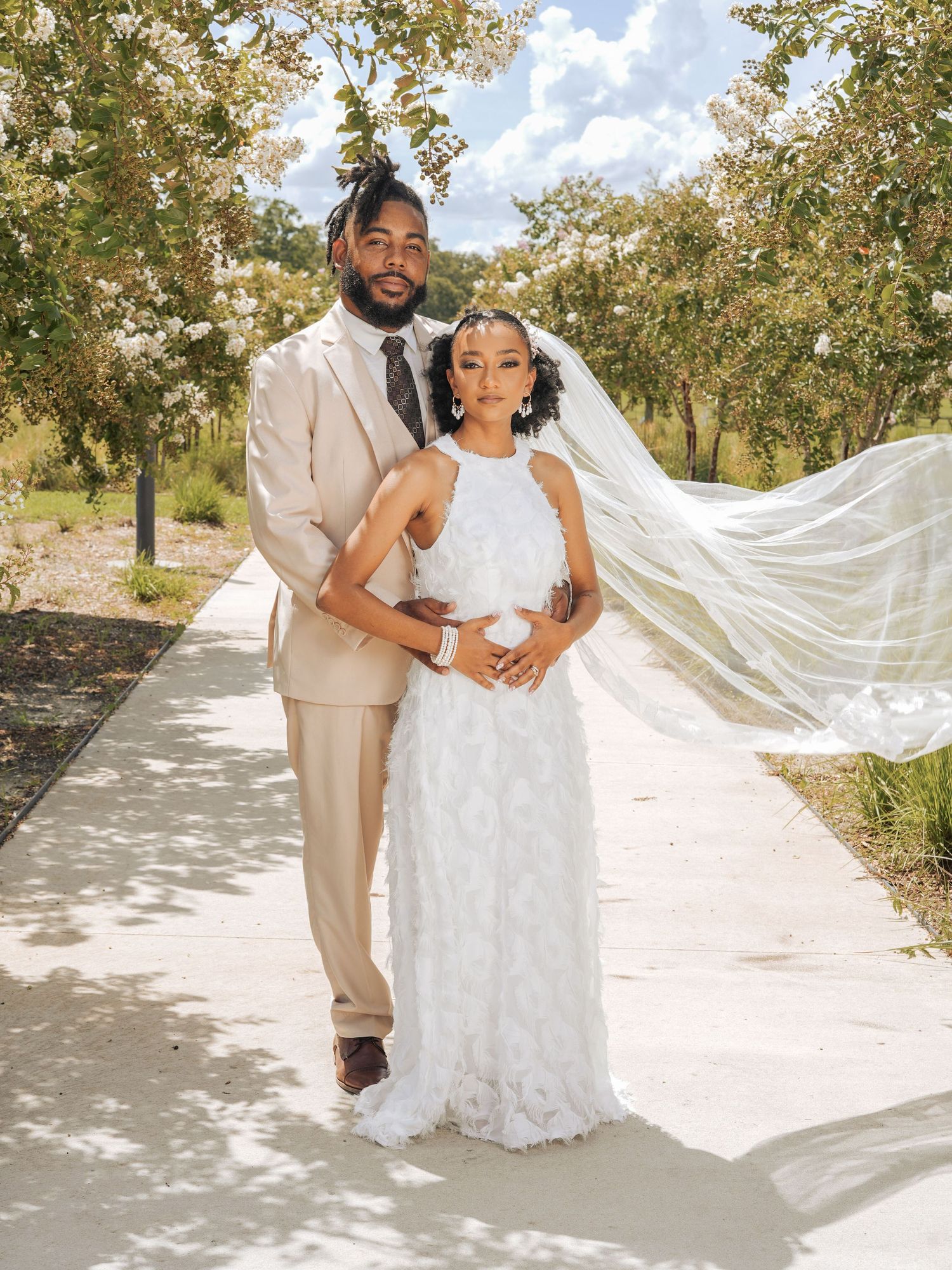 Newlyweds pose on sunny path with bride's veil flowing in the breeze.