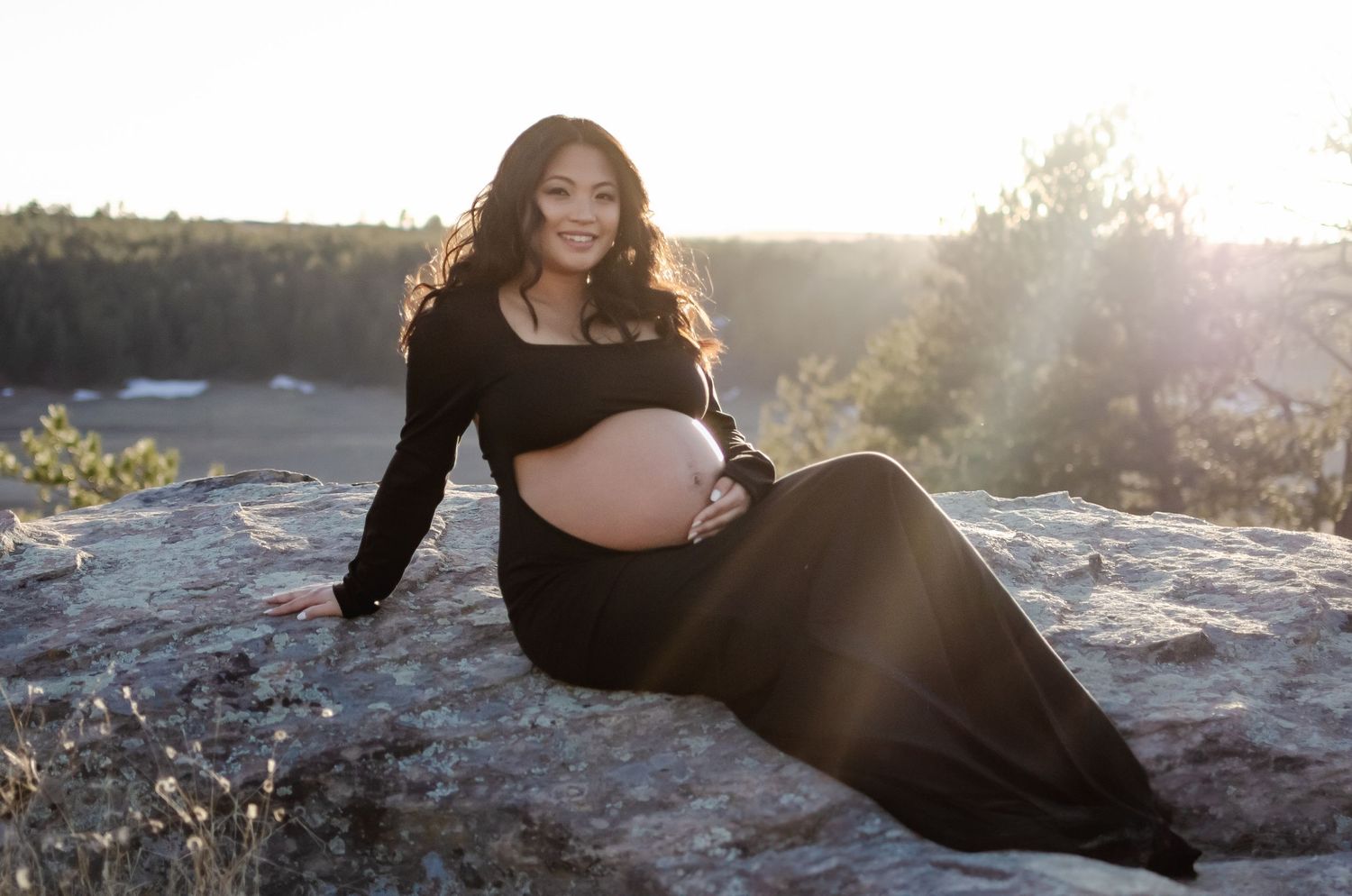 Pregnant woman in black dress poses on rocks during golden hour sunset.