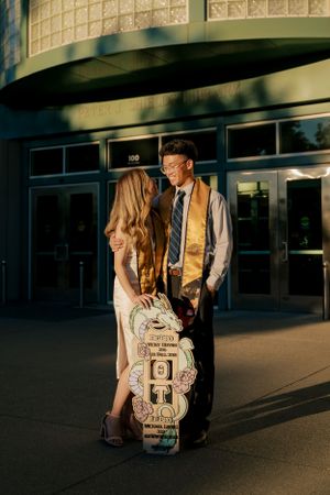 A couple stands together at sunset outside a building entrance with golden light and a decorated backpack.