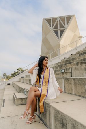 Graduate in white kimono and purple sash poses on concrete steps of modern building during graduation photoshoot.