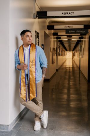 Graduate in blue shirt wearing gold graduation stole leans against wall in empty school hallway.