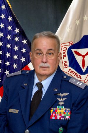 Military officer in Air Force uniform poses with American flag and Civil Air Patrol emblem.