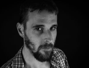 Dramatic black and white close-up portrait of person with beard wearing plaid shirt.