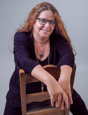 Professional portrait of someone wearing glasses and a dark sweater while seated casually on a wooden chair.