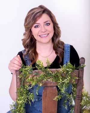 Person in denim overalls and black top poses with a rustic wooden ladder decorated with green foliage.