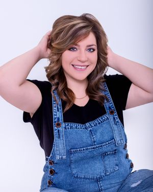 Smiling woman in denim overalls and black shirt poses with hands in hair against light background.