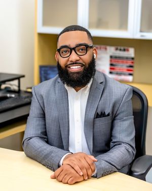Professional in a gray suit and glasses sits at a desk in an office setting with bookshelves in the background.