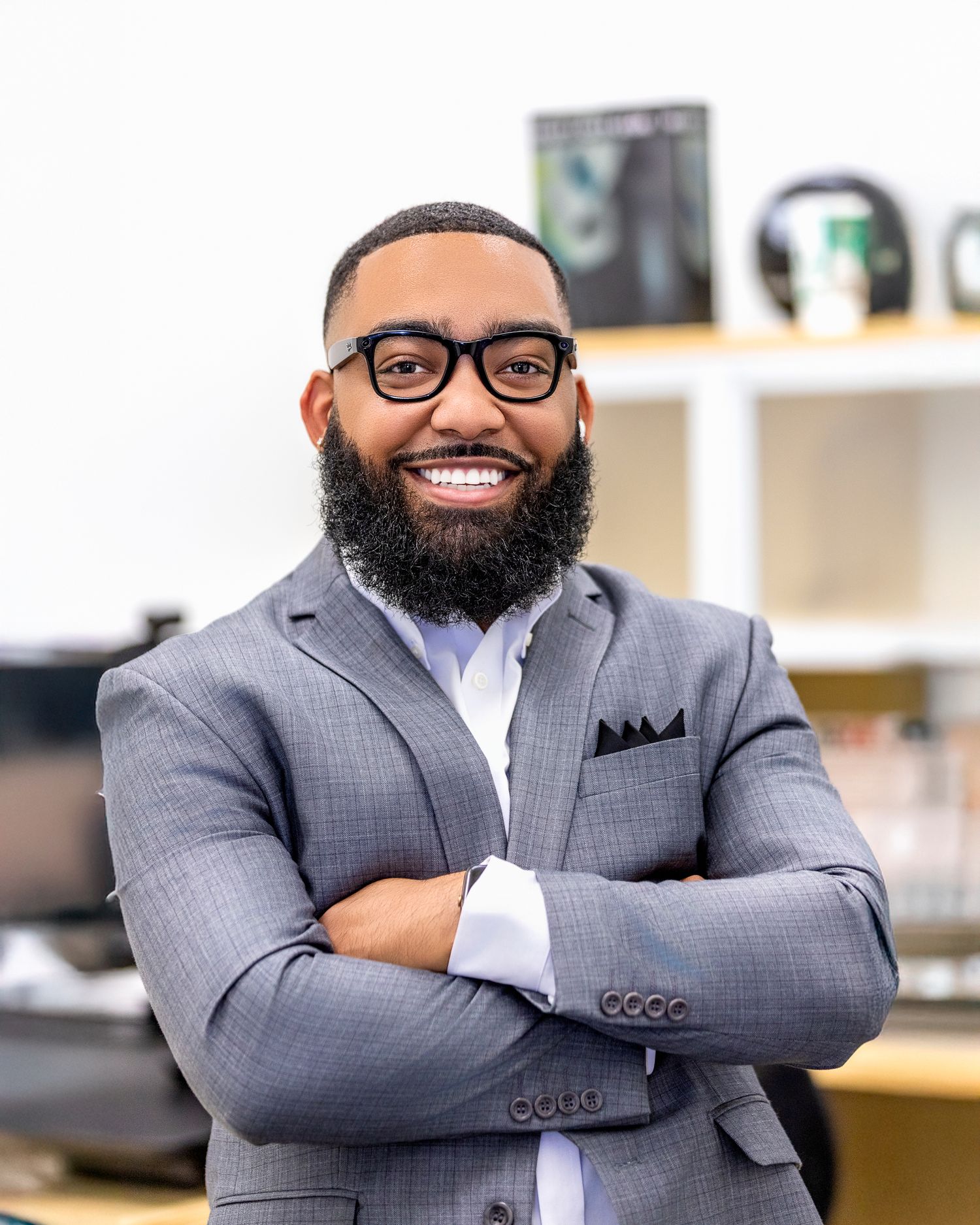 Professional wearing a gray suit and glasses poses confidently with crossed arms in an office setting.