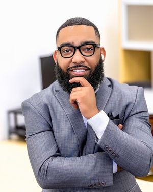 Professional in a grey suit and glasses sits thoughtfully at desk in office setting.