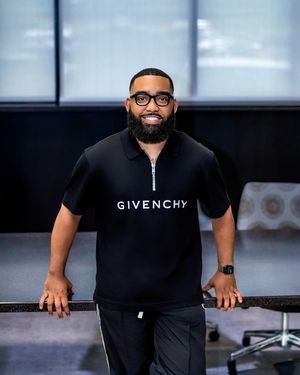 A person wearing glasses and a black Givenchy short-sleeved shirt smiles while leaning forward against a surface.