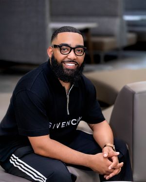 A person in a black t-shirt and glasses sits casually while smiling warmly at the camera.