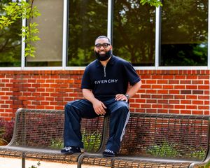 A person wearing a black Givenchy t-shirt sits on a metal bench outside a brick building.