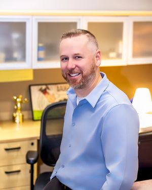 Professional wearing a light blue dress shirt smiles warmly in an office setting with natural lighting.