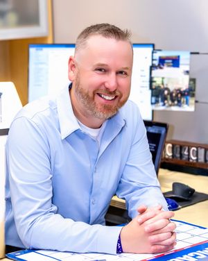 Professional in a light blue button-down shirt smiles while working at a modern office desk with documents.