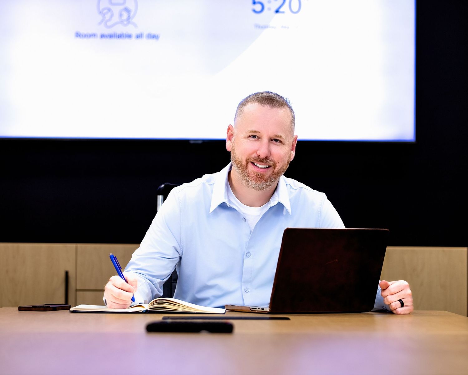 Professional wearing a light blue shirt sits at a desk in a conference room with a screen display visible behind them.