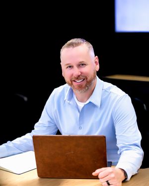 A person in a light blue dress shirt smiles while sitting at a desk in a professional office setting.