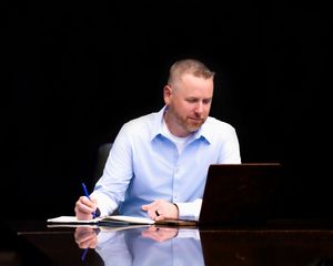 Professional in light blue shirt working at desk with laptop and documents in dark office setting.