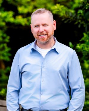 A person wearing a light blue button-down shirt smiles while posing outdoors with green foliage in the background.