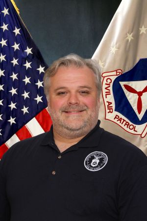 Official portrait of Civil Air Patrol member in black polo shirt with American and CAP flags in background.