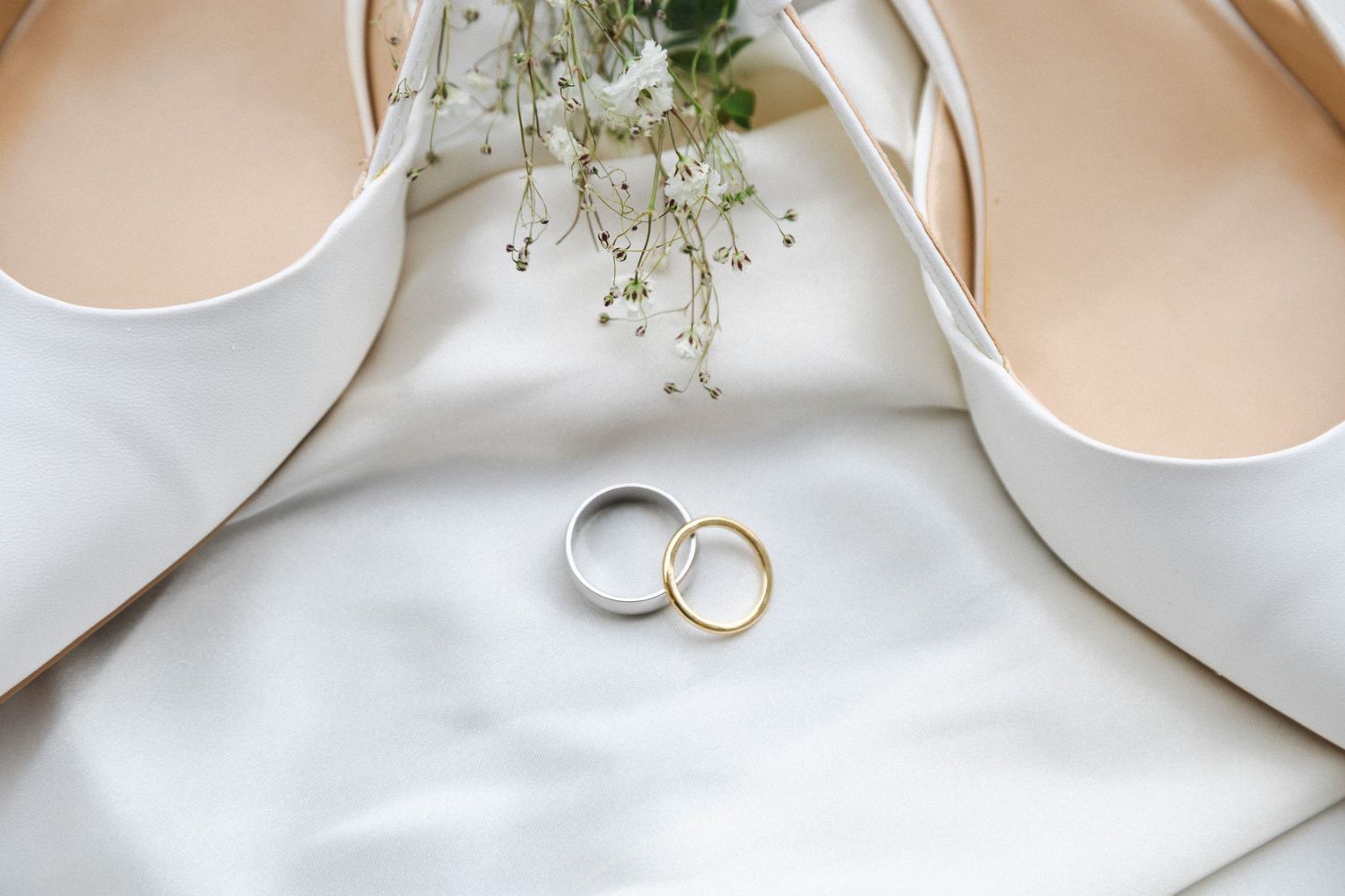 Wedding rings and delicate floral decorations arranged on white fabric with soft natural lighting.