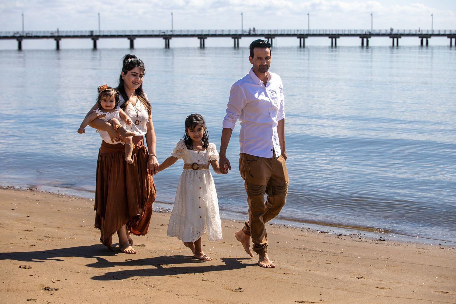 A family walks along a scenic beach with a pier visible in the background.
