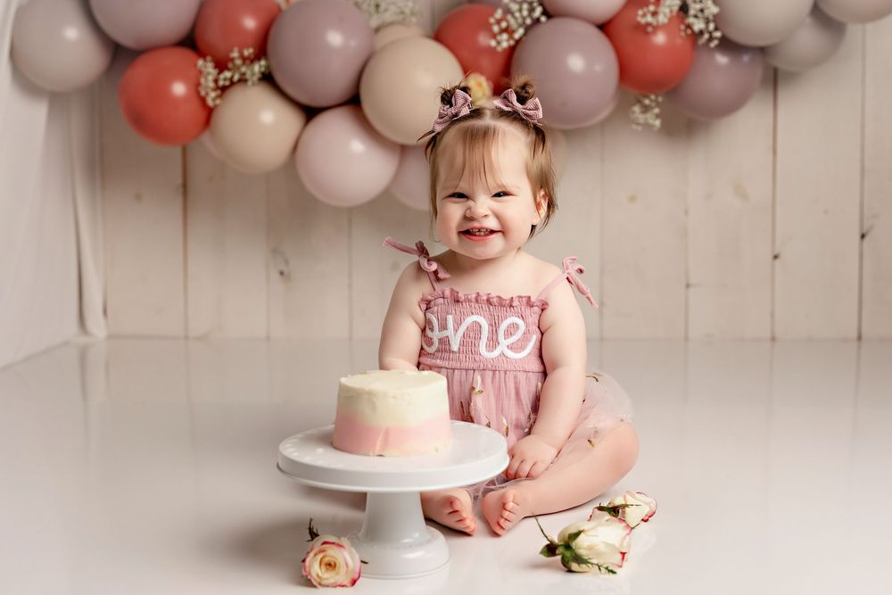 First birthday celebration setup with pink and gray balloon arch, white cake on stand, and flowers against white backdrop.  Kendall Kuss
