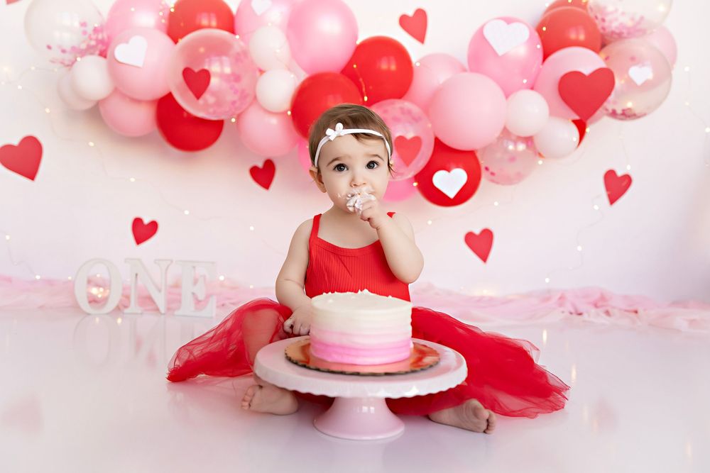 Pink and red balloon decorations with hearts create a festive backdrop for a first birthday cake smash photoshoot.  Kendall Kuss Photography