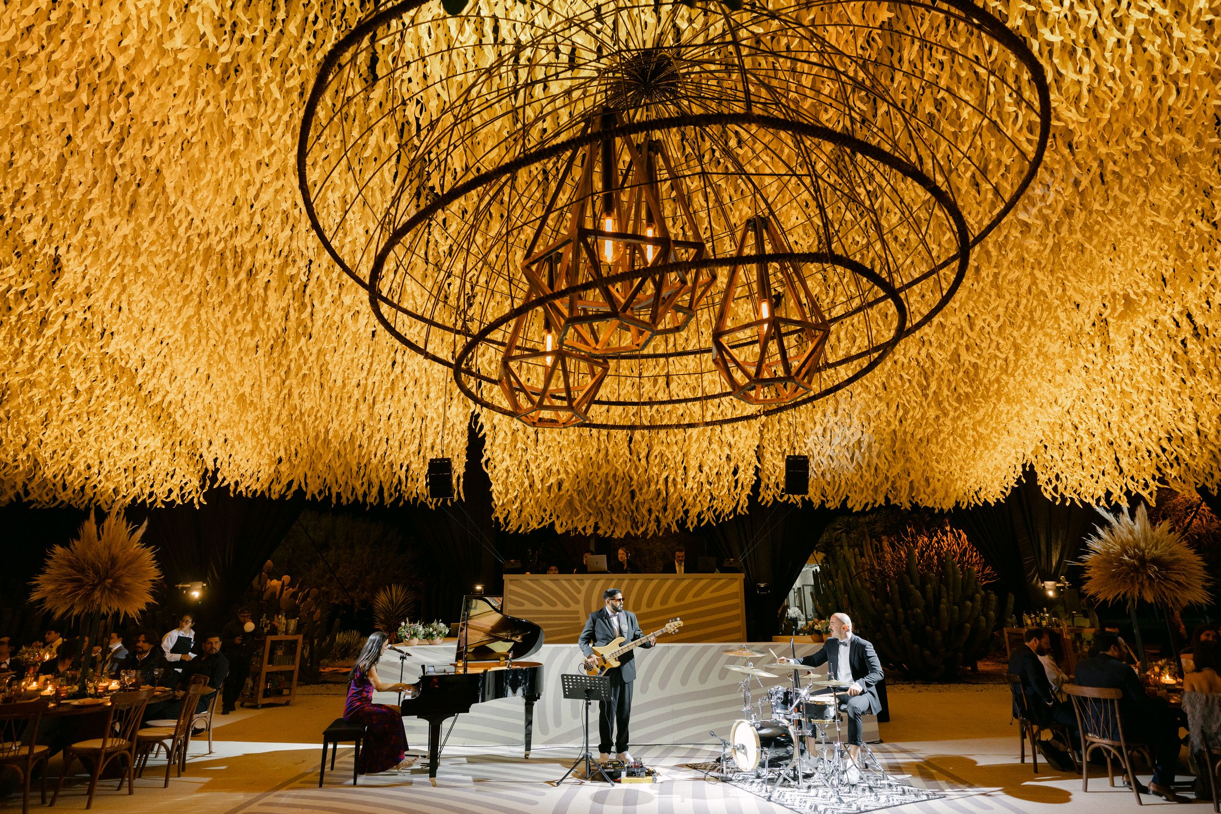 Musicians perform on a concert stage beneath a dramatic circular chandelier installation with golden ambient lighting.