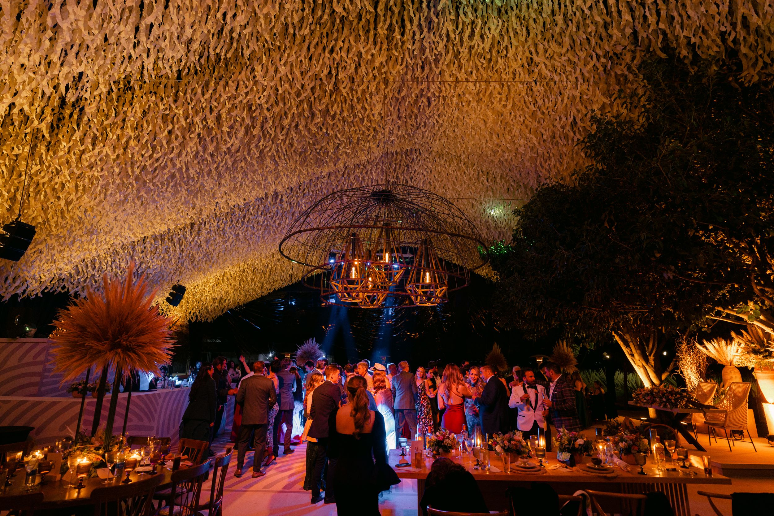 Wedding guests dance and celebrate under a colorful illuminated thatched roof at a tropical evening reception.