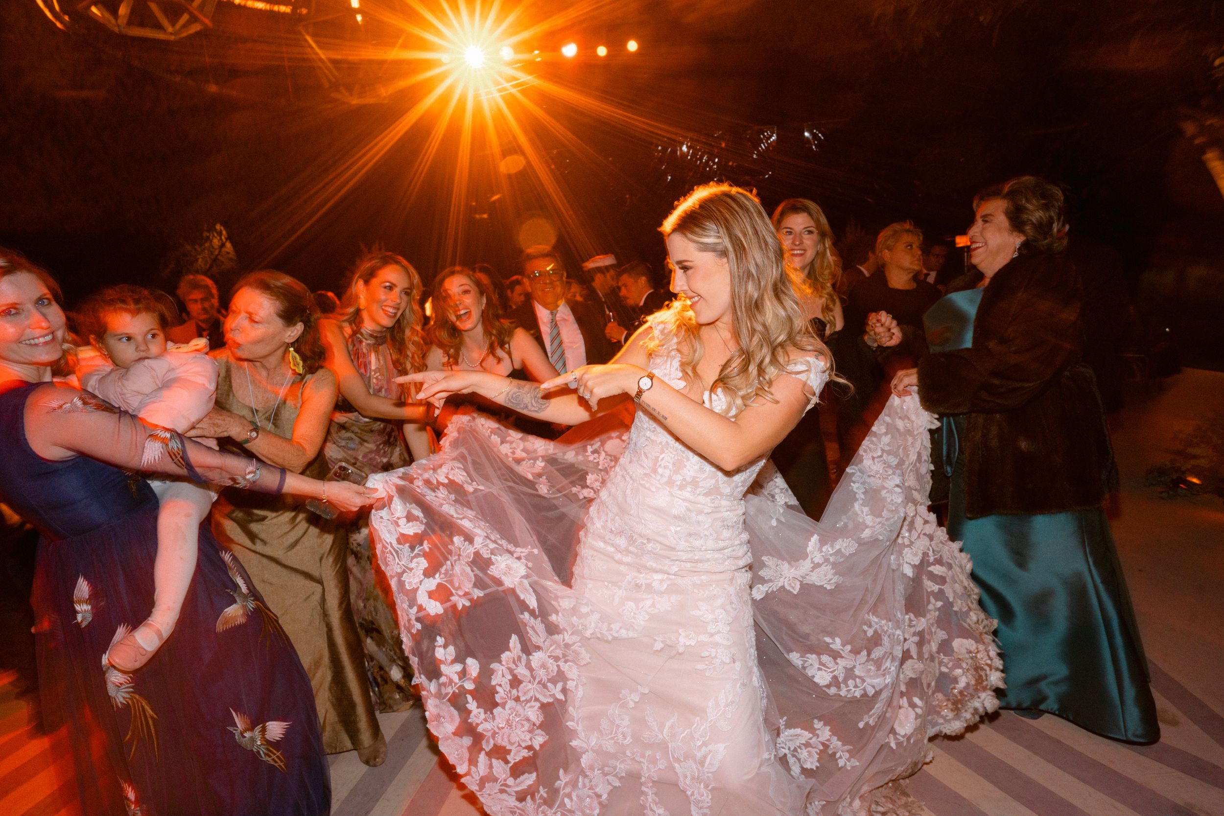 A bride in a lace wedding dress dancing with guests at a dimly lit reception under warm lighting, beautifully captured by Alejandro Cayetano