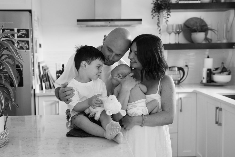 Black and white family photo of parents with newborn baby and young son in a bright modern kitchen.