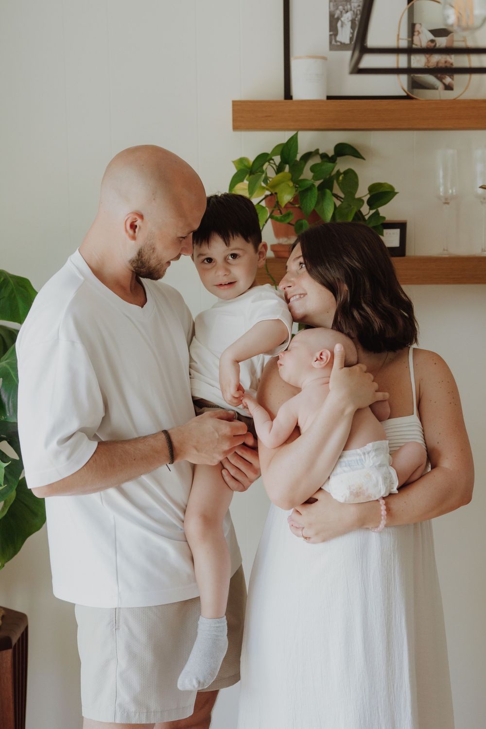 Happy family of four in white outfits, parents holding toddler and newborn baby indoors with green plants.