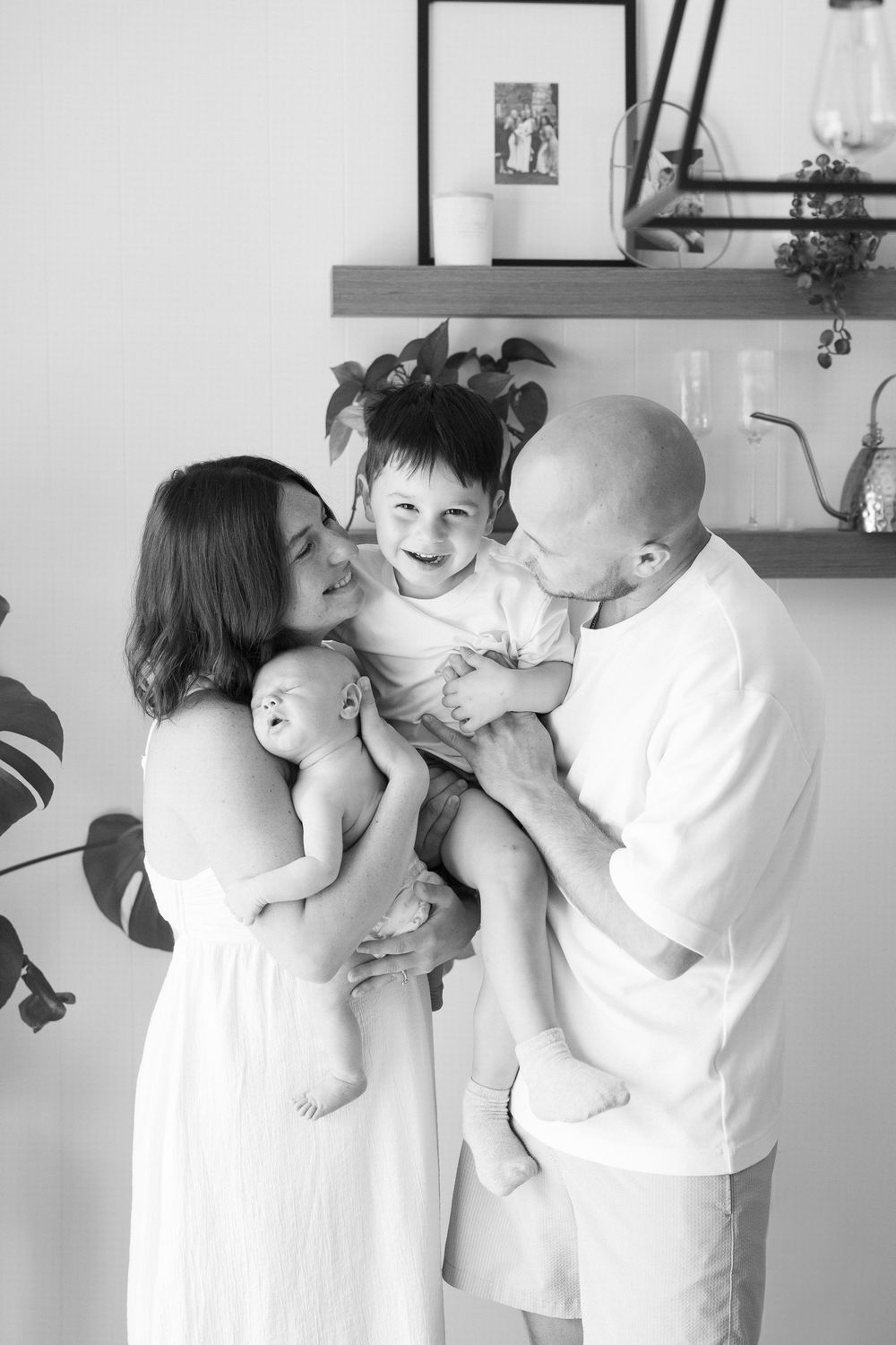 Black and white family photo of parents kissing toddler while mother holds newborn baby indoors.