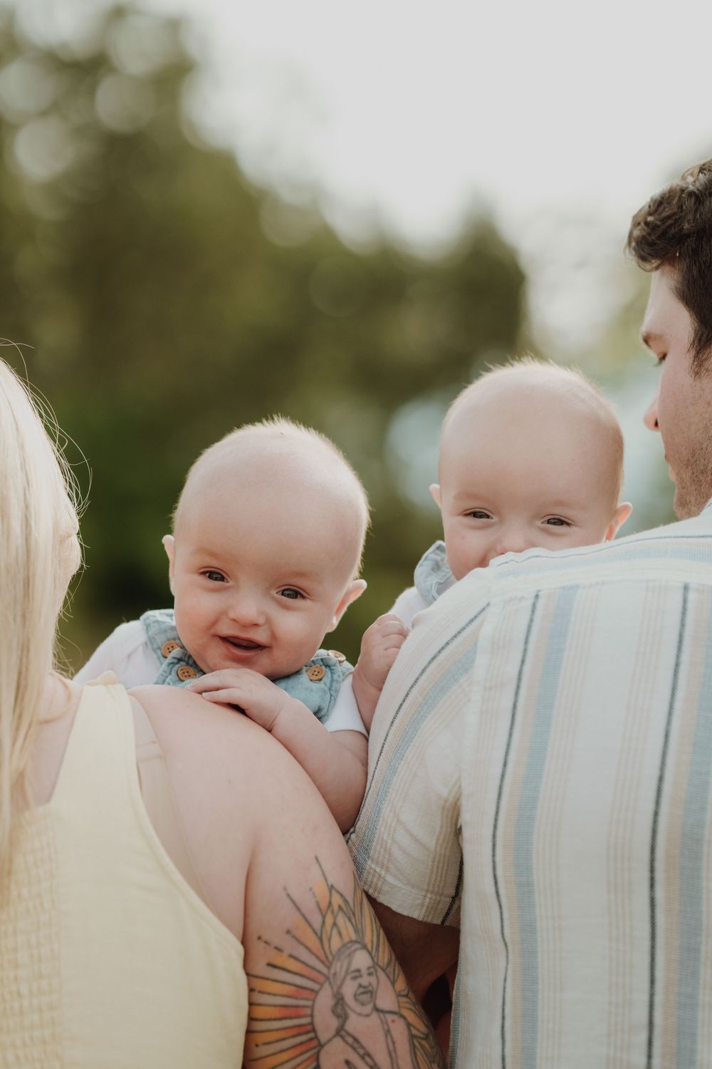 Two smiling babies held by their parents outdoors, with a tattooed arm visible and soft green bokeh background in Victoria BC.