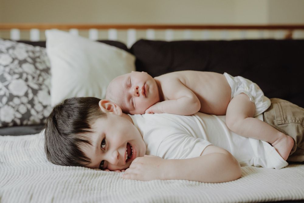 Young boy lying on bed smiling with newborn baby sibling resting on his back, sweet sibling bond moment.