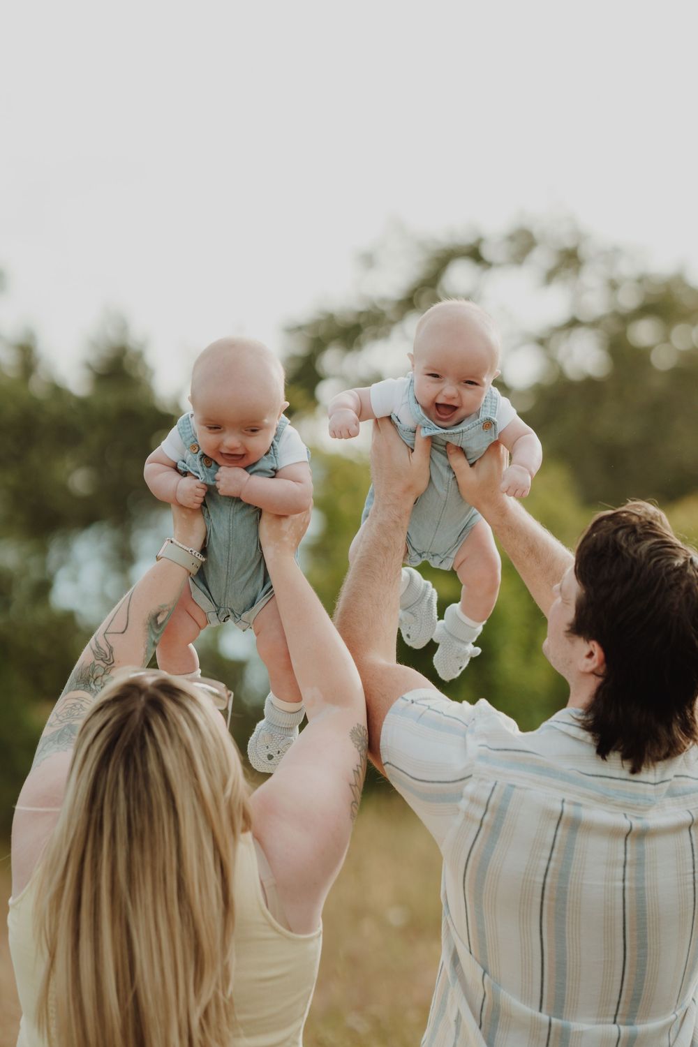 Joyful parents lifting their twin babies in matching outfits high in the air in a sunny outdoor field in Victoria BC Vancouver Island.