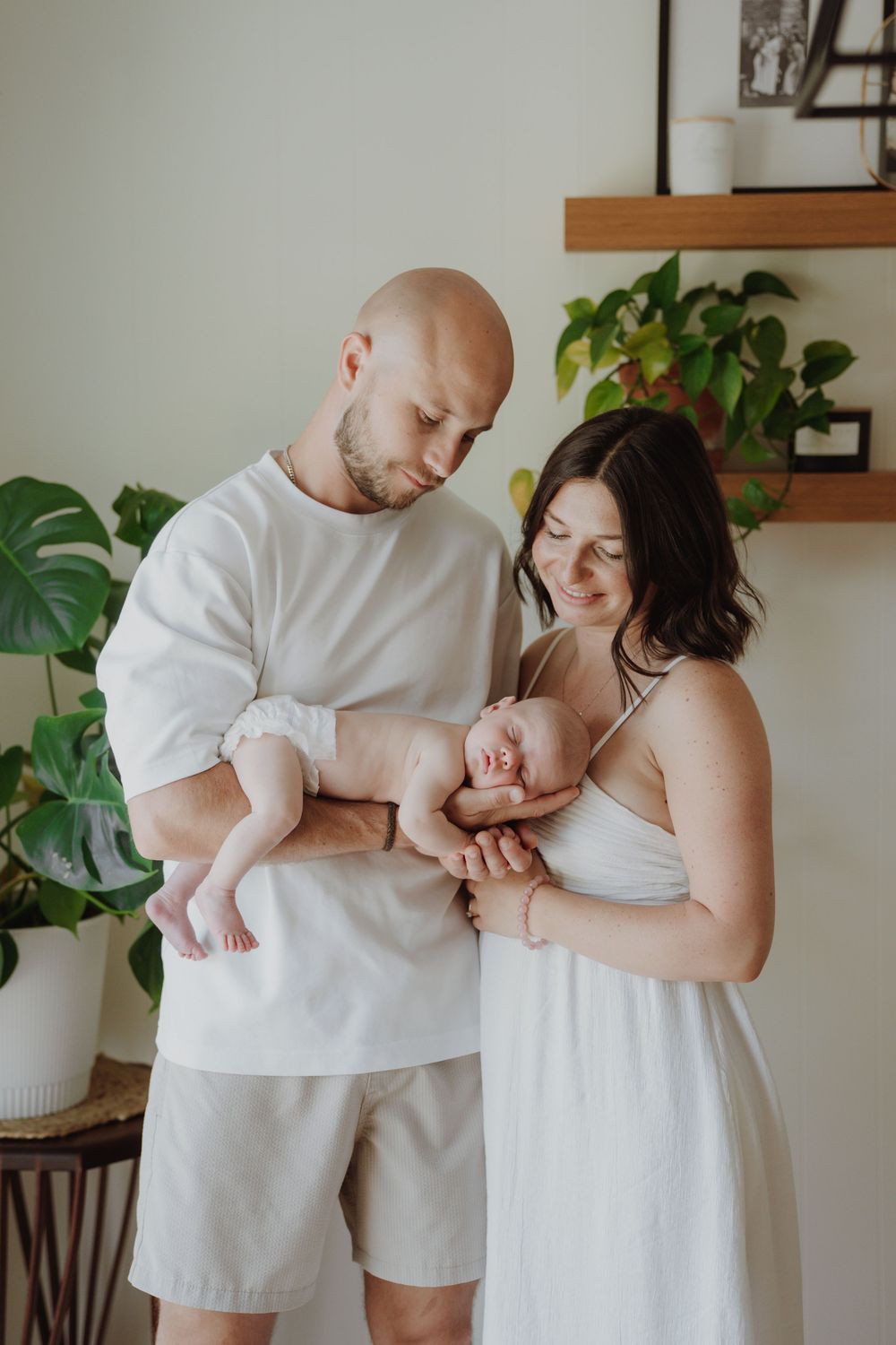 A bald father and dark-haired mother in white outfits tenderly hold their newborn baby indoors with green plants.