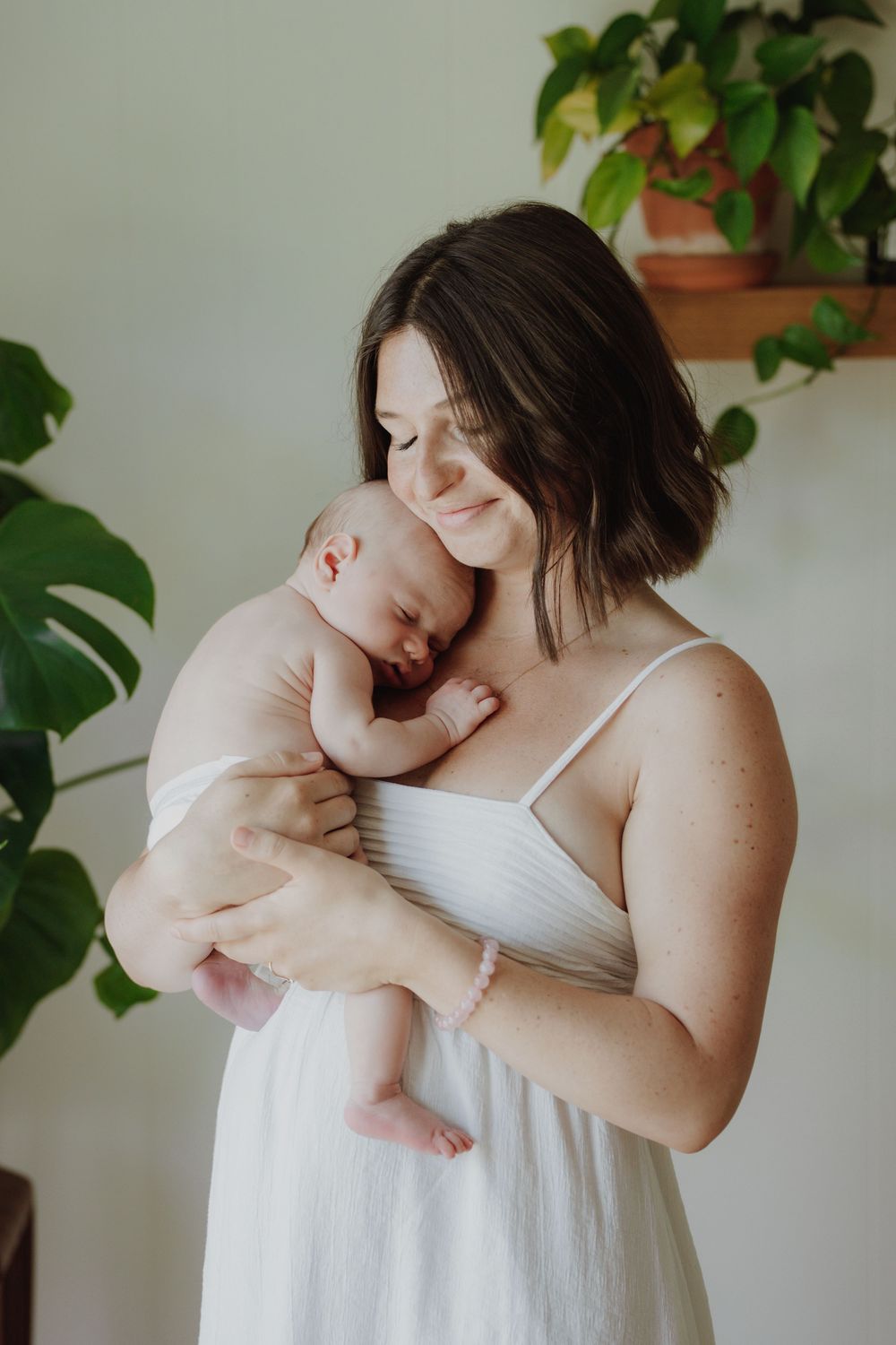 A mother in a white dress tenderly holds her sleeping newborn baby, surrounded by lush green houseplants.