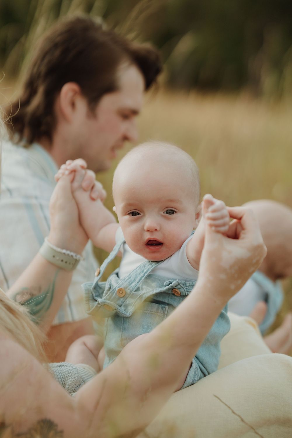 Adorable baby in denim overalls held by parents while sitting in a golden grass field during family photos in Victoria BC Vancouver Island.