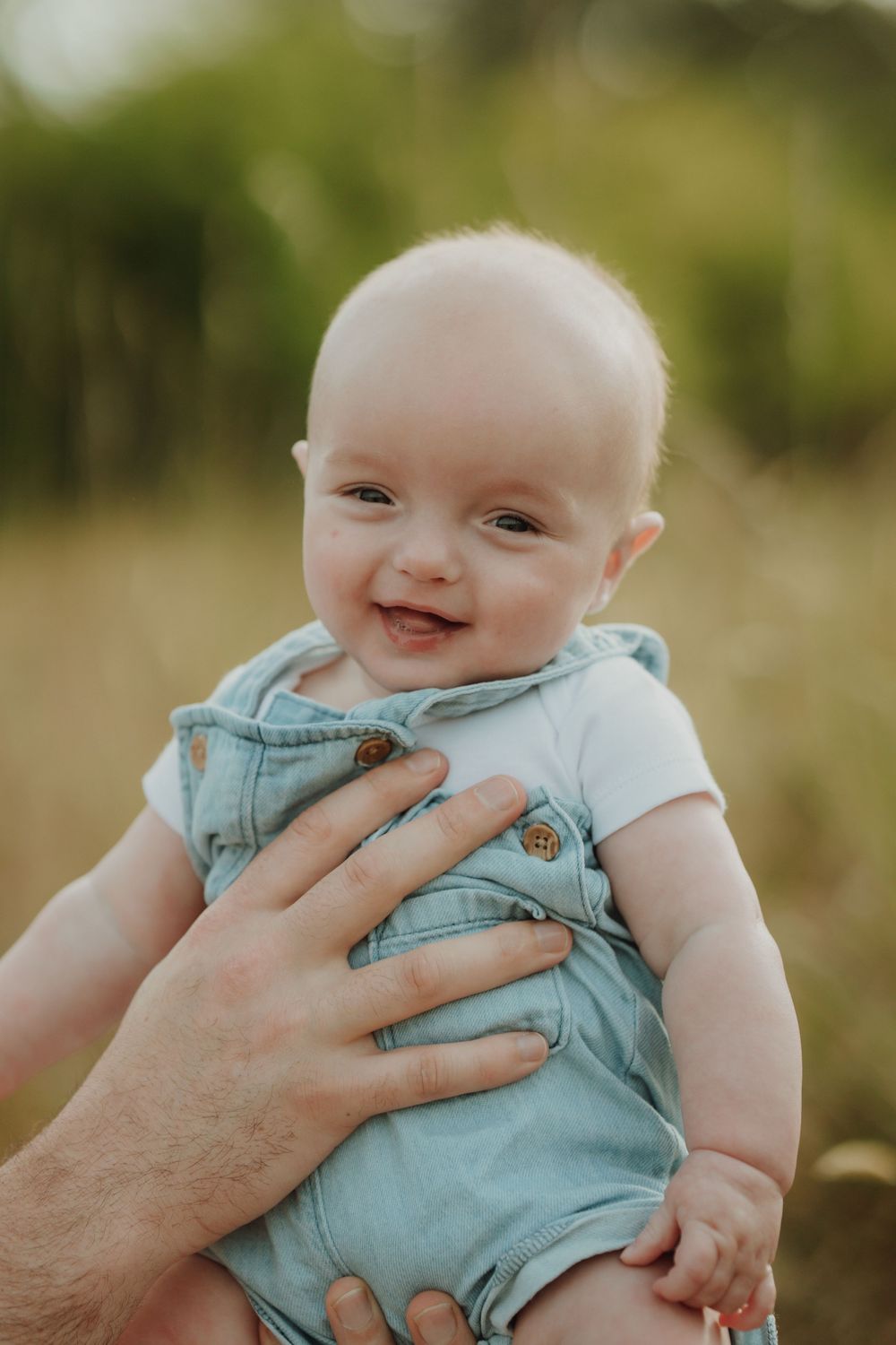 Happy smiling baby in denim overalls being held outdoors in a natural field setting during a family session. In Victoria, BC.