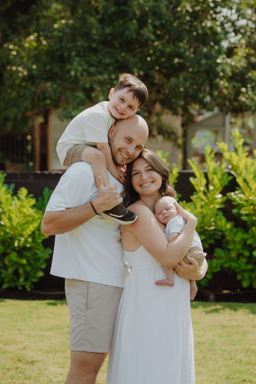 Happy family of four posing outdoors, parents holding toddler and newborn baby in a lush green garden.
