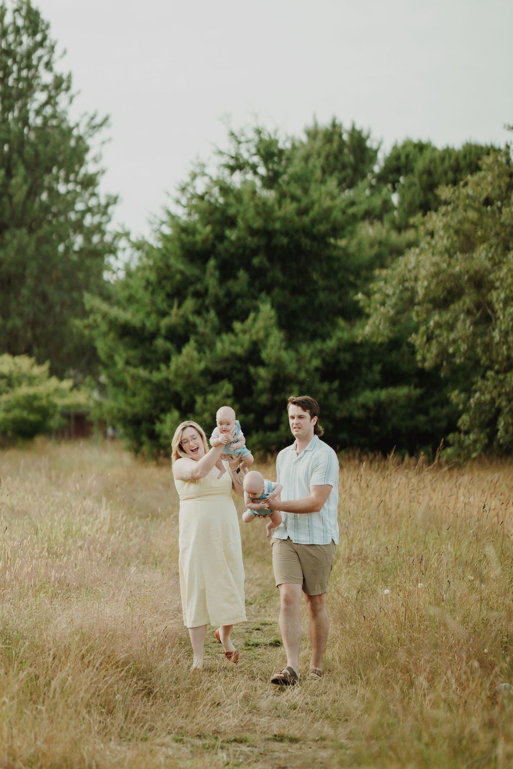 Young family with twin babies walking through a golden meadow surrounded by tall green trees in soft light. In Victoria, BC.