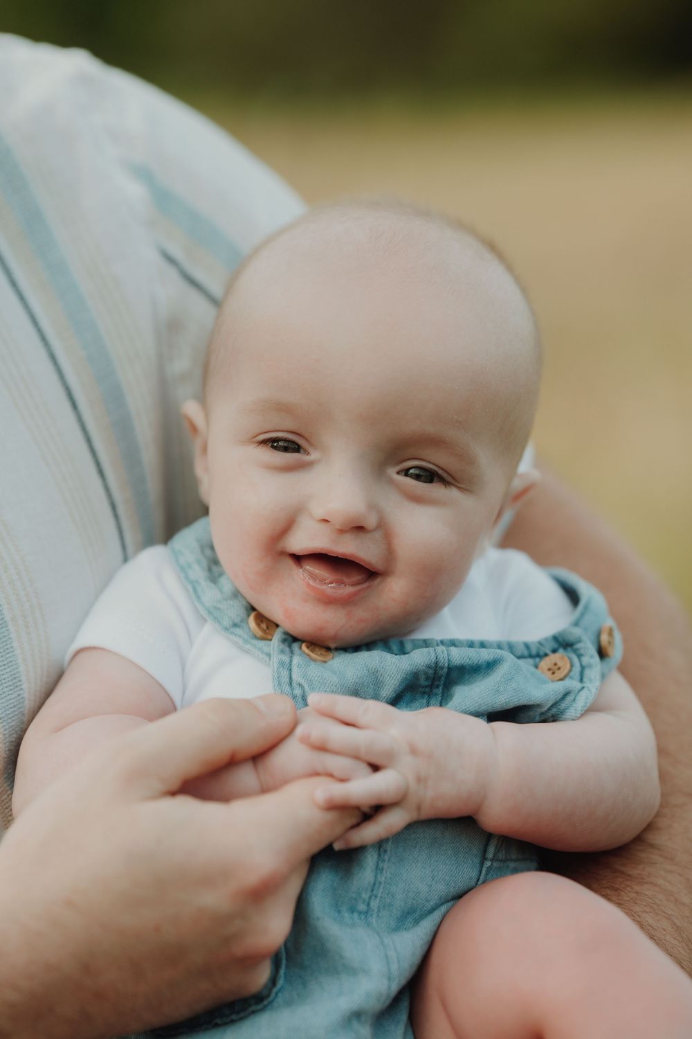 Cheerful baby in denim overalls smiling brightly while cradled in a parents arm during an outdoor photo session. In Victoria, BC, Vancouver