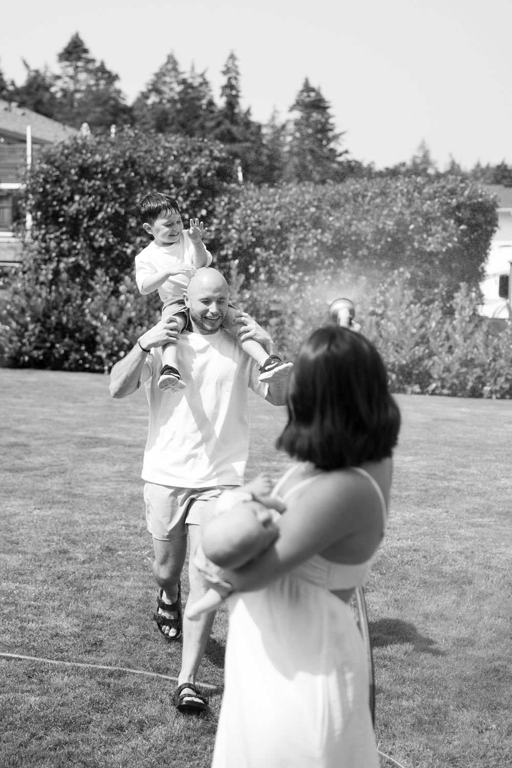Black and white photo of a man with child on shoulders interacting with a woman outdoors on a sunny day.