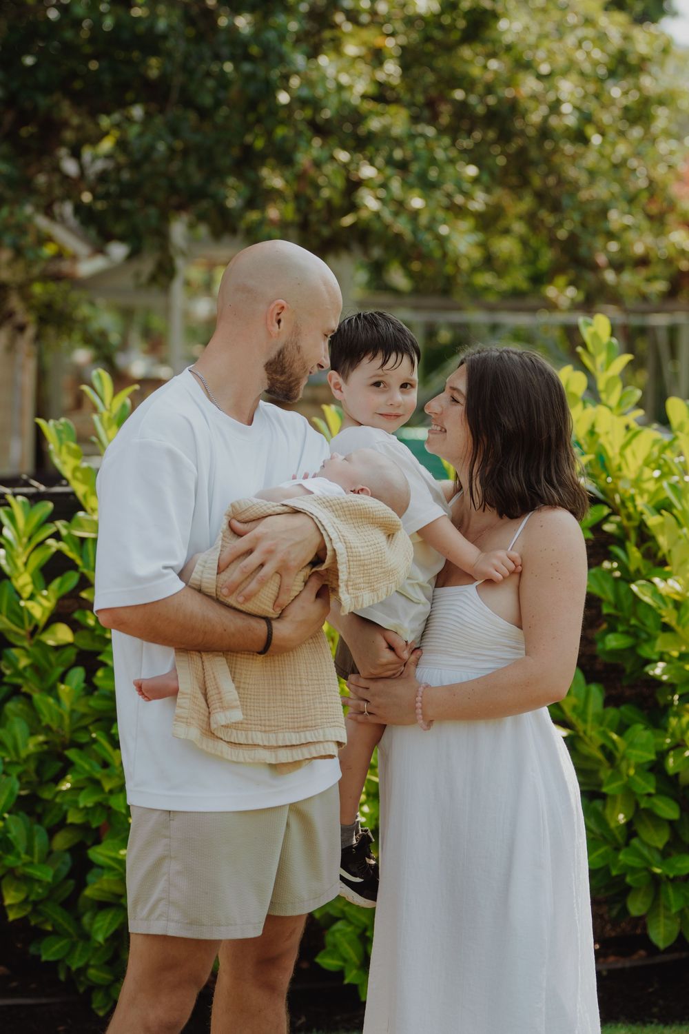 A young family with a toddler and newborn baby poses outdoors surrounded by lush green tropical foliage.