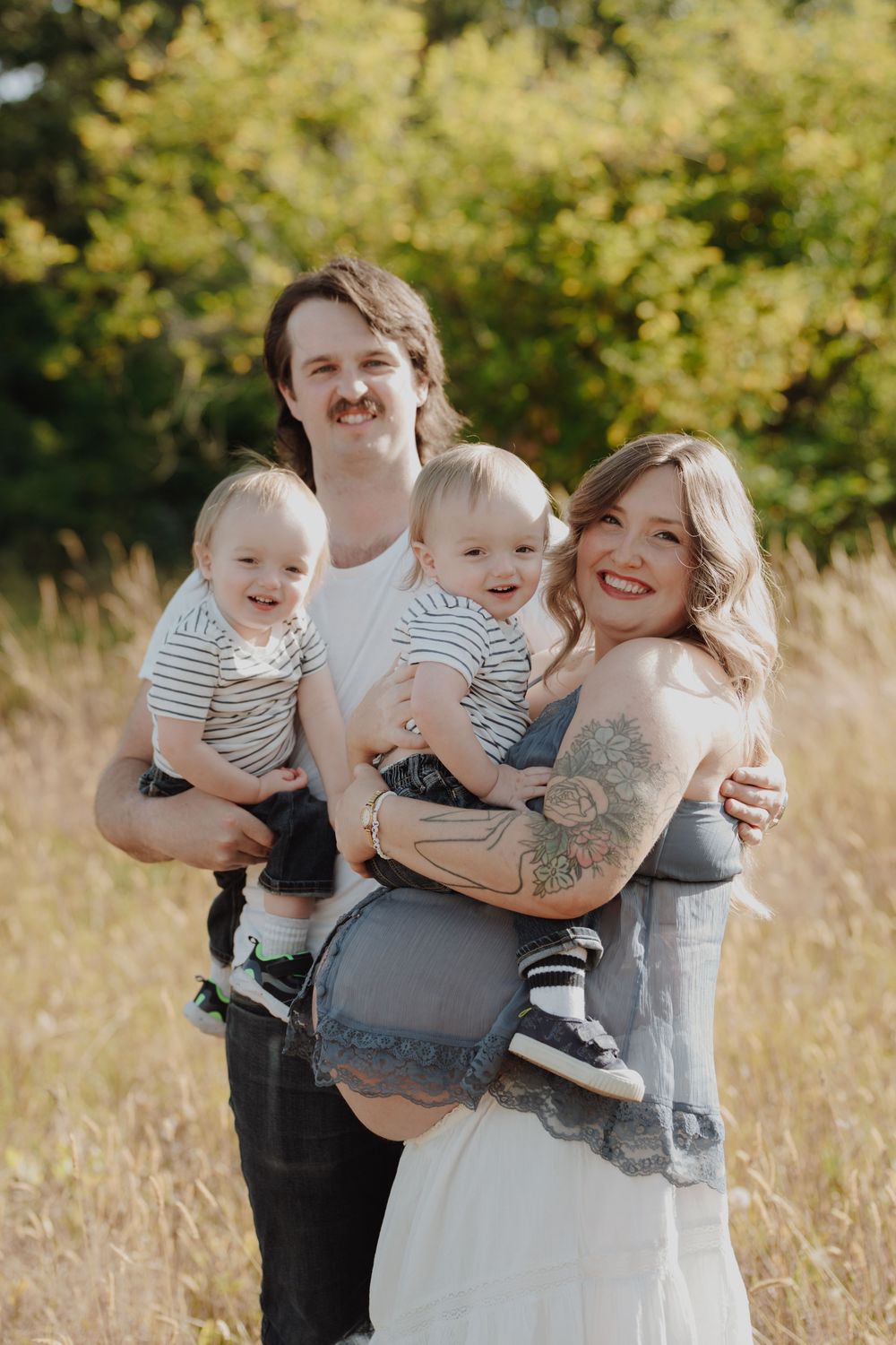 A smiling couple holds two babies in a sunny field, the woman with tattoos wearing a gray dress and denim jacket.