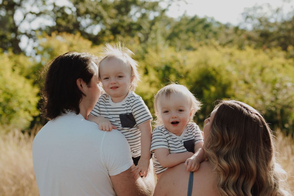 Parents holding twin babies in matching striped outfits outdoors in a sunny field with green trees.